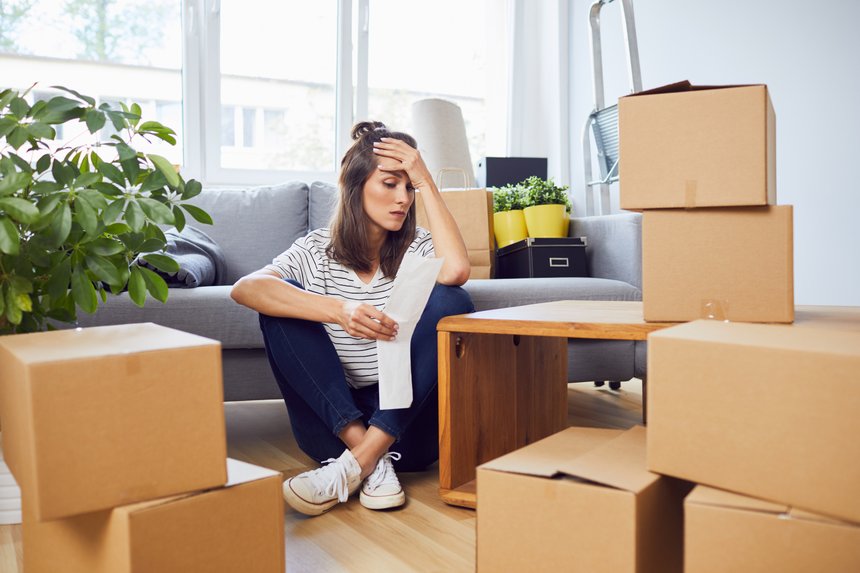 Worried young woman looking at bill sitting on floor in new apartment after moving in