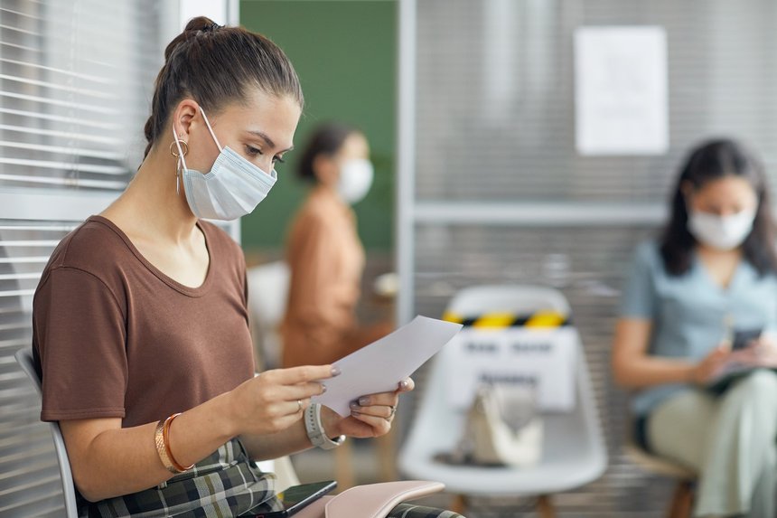 Side view portrait of young woman wearing mask and holding document while waiting in line in office, copy space