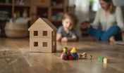 Wooden house model on the floor, a family playing with toys in a blurred living room background.